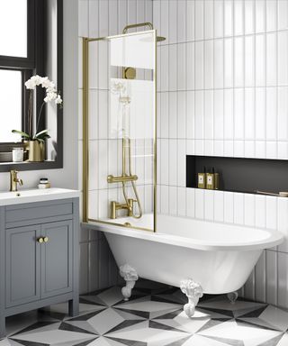 bathroom with roll top bath, slim gold shower screen to side, gold wall mounted shower riser, black and white tiled floor, white tiled wall and black framed window with grey vanity unit and small sink below