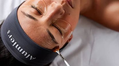 A woman getting a facial while wearing a headband that says AlumierMD.