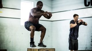 A man wearing shorts and cross training shoes jumps onto a box during an exercise class. His legs are bent and his arms are in front of him, as he catches his balance. Behind him we see another man exercising and a white-washed brick wall.