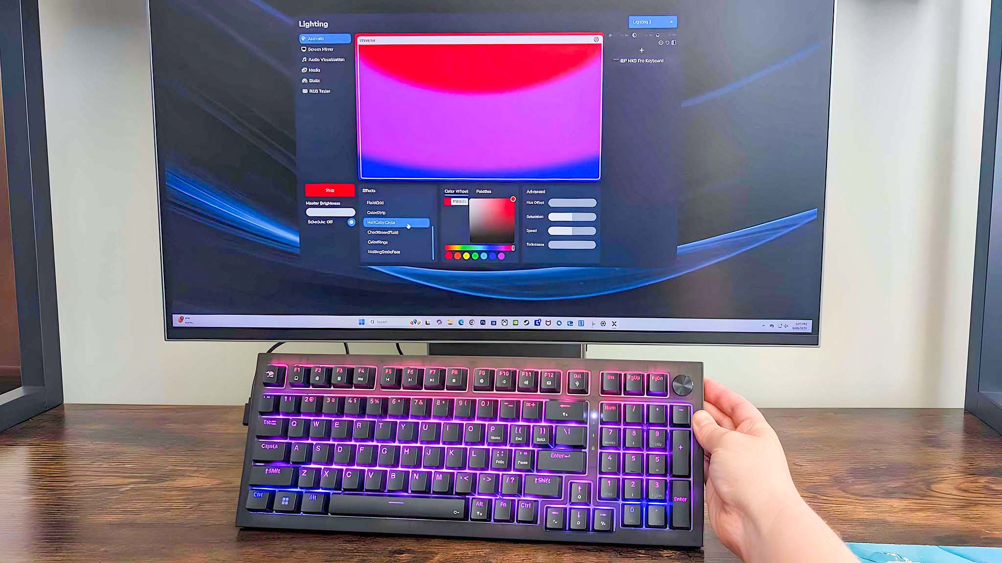 A hand holding the iBuyPower MK9 Pro RGB Mechanical Keyboard up and showing that the keyboard's rainbow RGB effect matches what's shown on the computer's screen in the back.