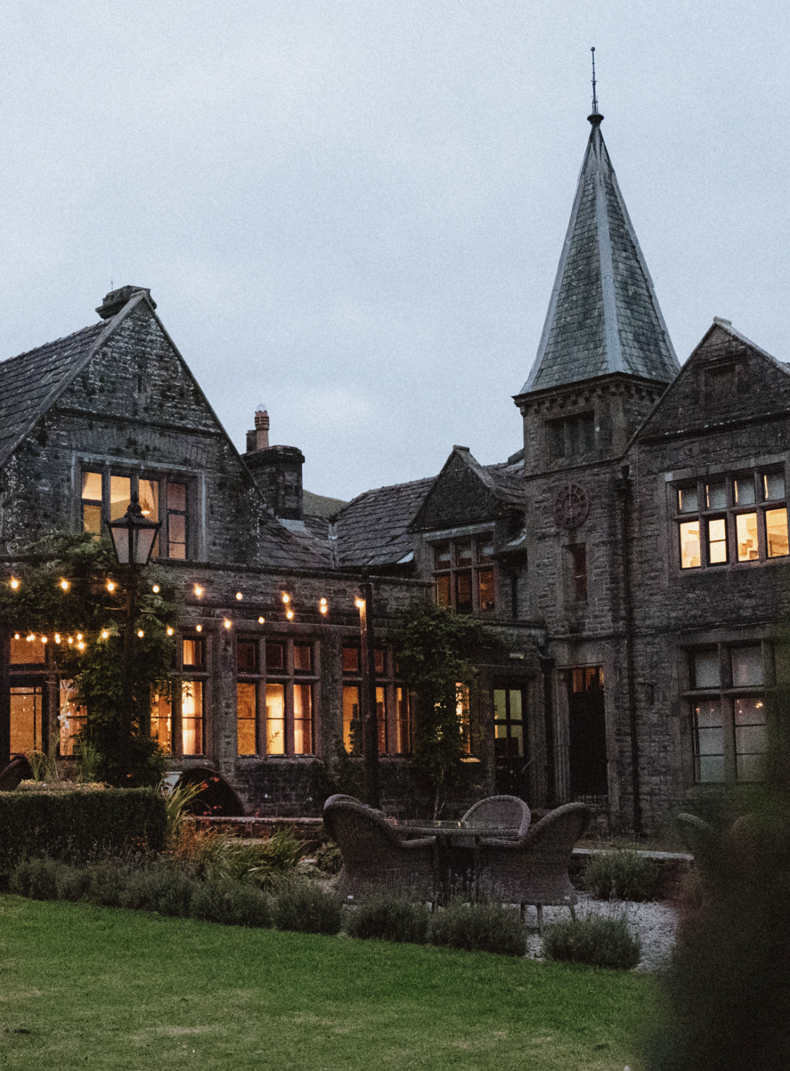 The brick exterior of a historical country lodge immersed in greenery is captured at dusk as the warm lights of its interiors come on, with ivy running over its outside walls.