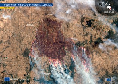 A brown landscape is scarred dark red as streaks of orange flames are seen around the edges of the dark area with streams of smoke moving toward the bottom of the image