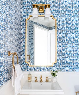 A blue bathroom with a gold mirror, white sink, and gold towel ring with a white towel