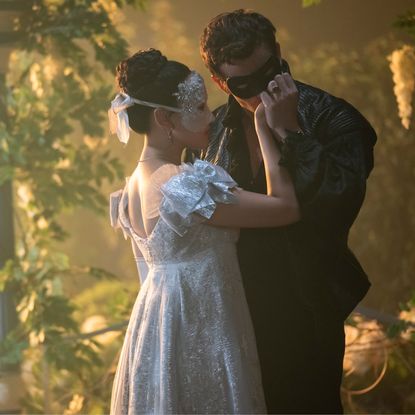 A masked man (Luke Thompson as Benedict Bridgerton) kisses a masked woman's (Yerin Ha as Sophie Baek) hand as they stand outdoors, in 'Bridgerton' season 4.