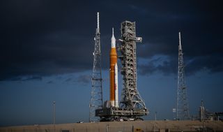 An orange rocket stands on a mound before a dark blue sky.