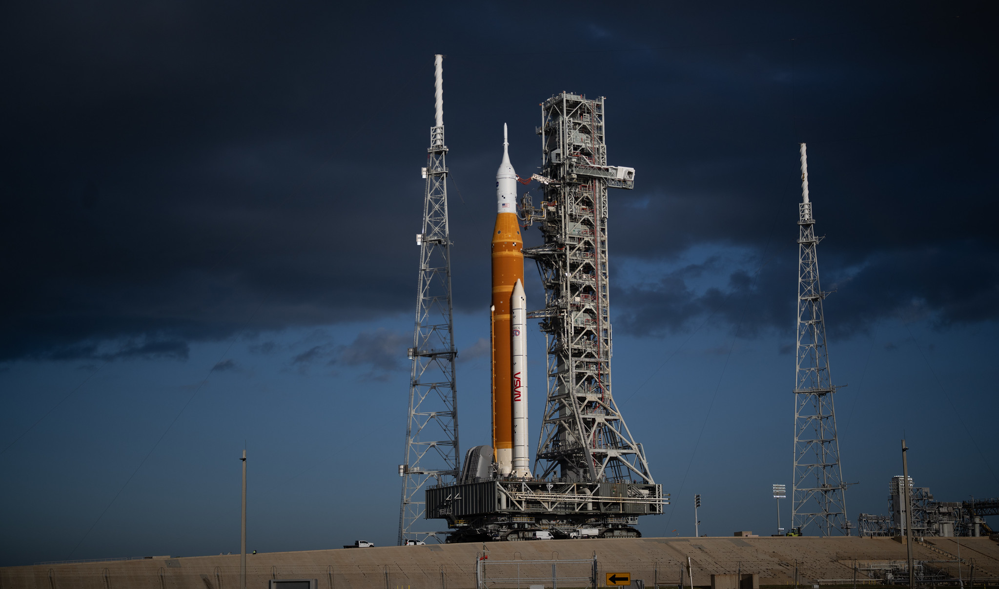 An orange rocket stands on a mound before a dark blue sky.