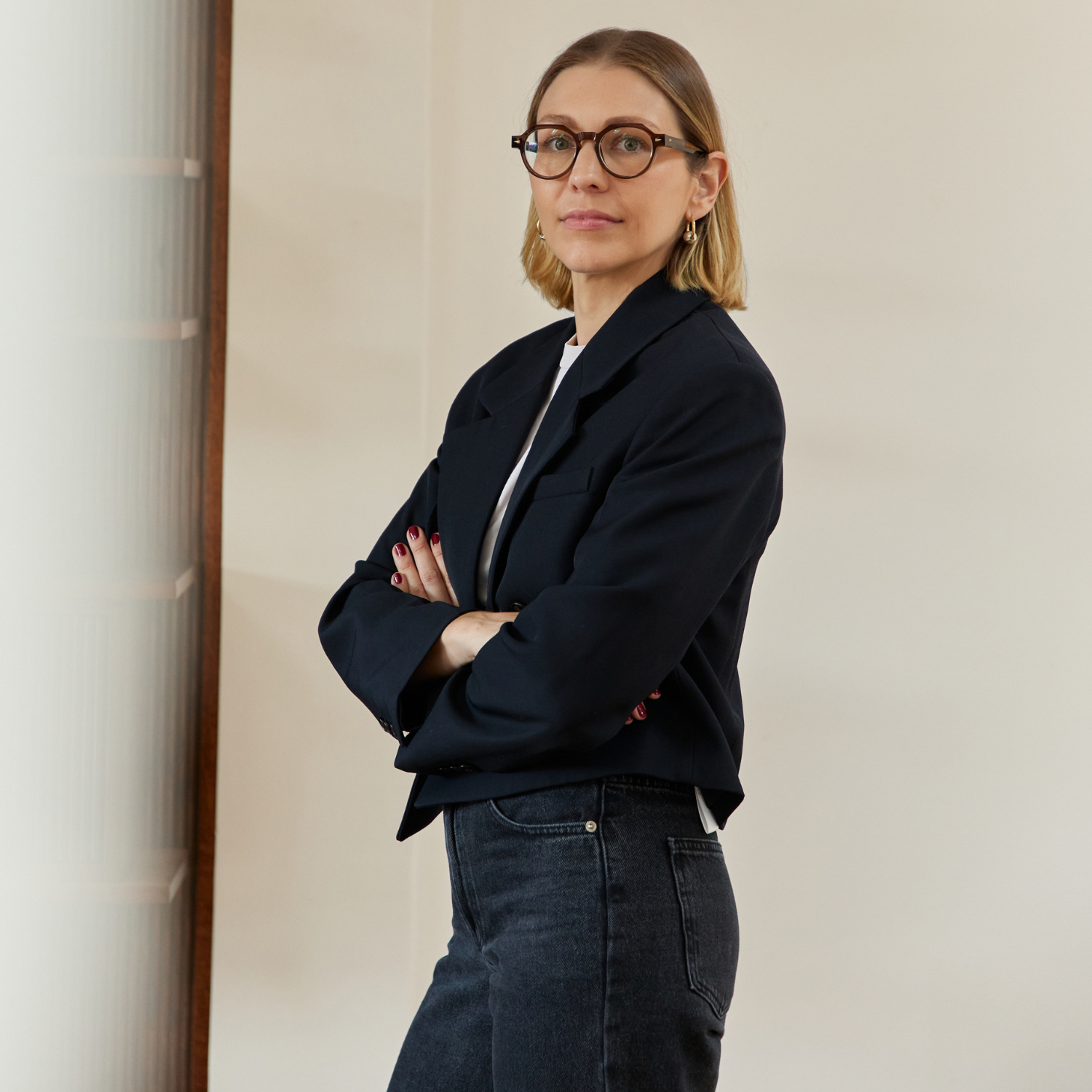 A woman with short hair and round glasses in a cropped blazer and jeans is posing with her arms crossed.