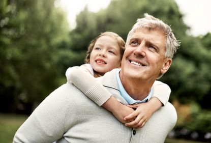 A young granddaughter gets a piggy back ride from her grandfather in the park. They are both smiling and looking left of the camera.