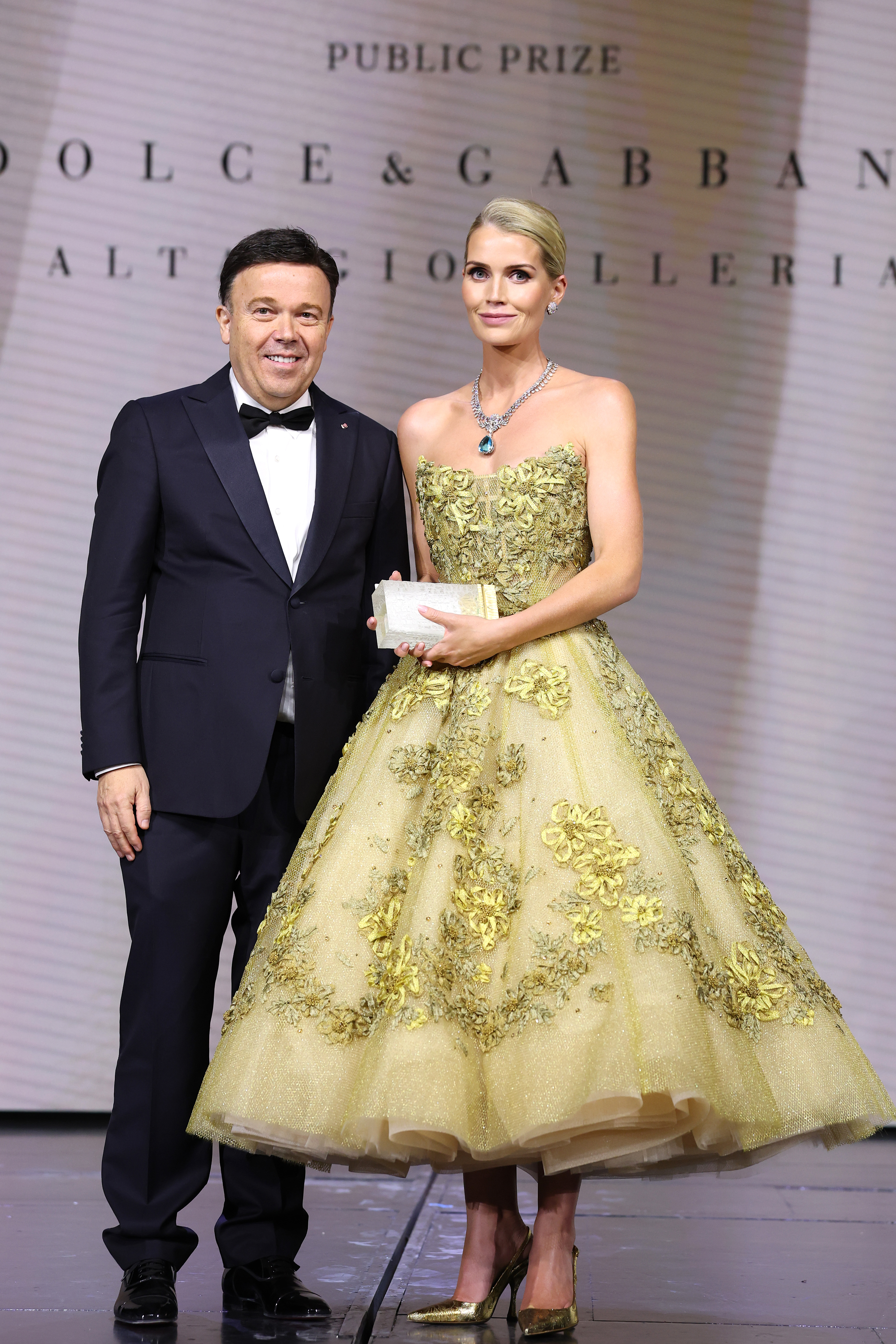 Kitty Spencer wearing a yellow tulle gown and a huge diamond necklace holding an award next to a man in a tuxedo on stage