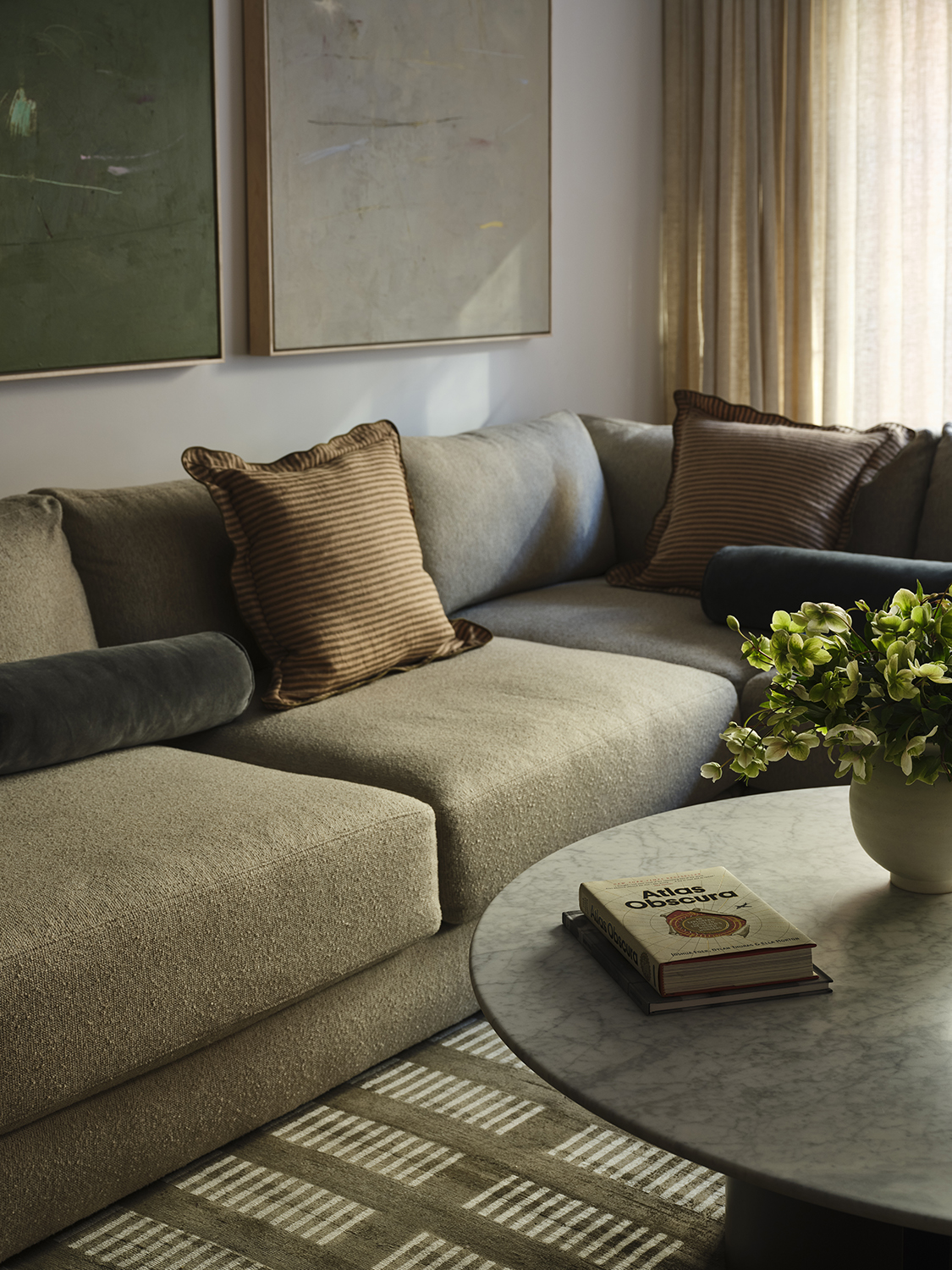 Close-up of a beige sectional sofa with a pillow on it and a marble, round coffee table in front of it. There is a patterned rug on the ground and two paintings on the wall.