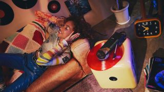 A woman lying on a sofa listening to a red vinyl record playing on the Audio-Technica Sound Burger AT-SB727. The picture also features a clock, posters, vinyl covers on the floor and vinyl records used as decorations on the wall.