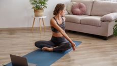 Woman performing a yoga pose sitting cross-legged on a yoga mat in front of a laptop in a living room