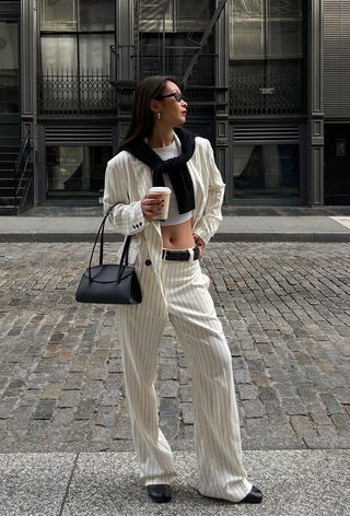 A woman wearing a white pinstripe blazer with matching trousers styled with a black cardigan tied around the shoulders, a white cropped tank top, a black belt, black ballet flats, and a black shoulder bag.