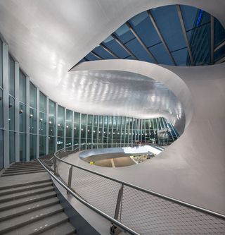 An inside view of Arnhem Station with a staircase going up along a curved glass wall.