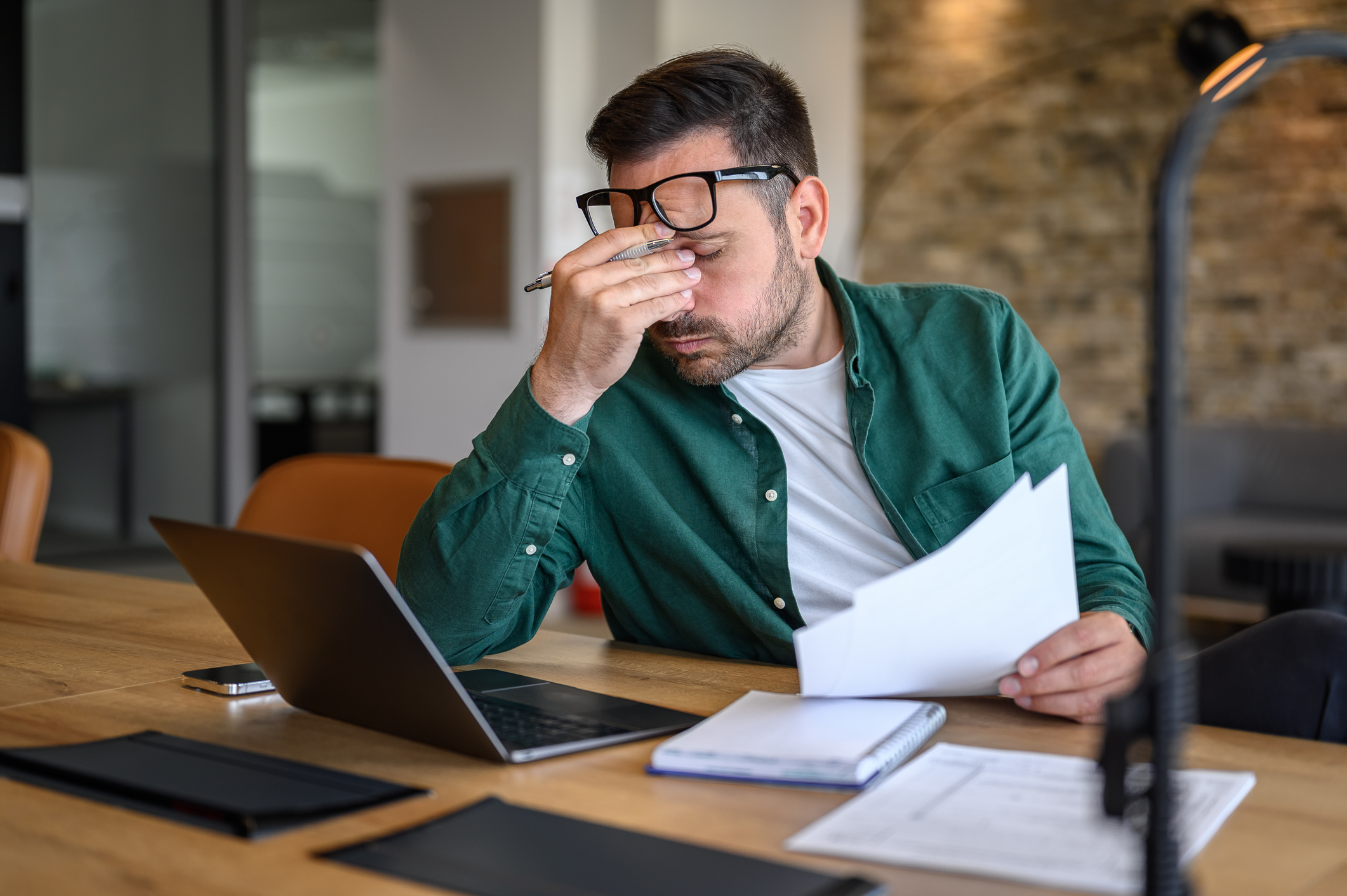 Anxious-looking financial advisor rubbing eyes while working over laptop and analyzing reports at desk