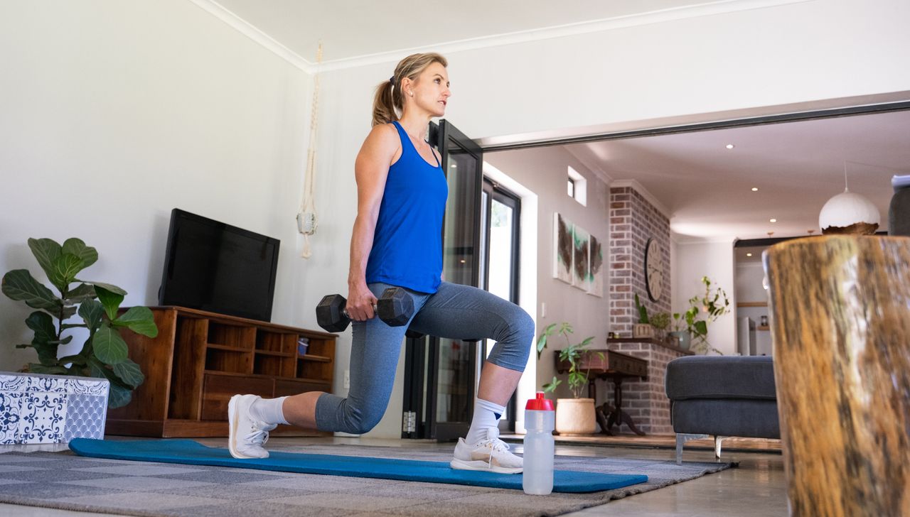 Woman performs lunge while holding dumbbells by her sides. One front is front of her and the other is behind her, and both her knees are bent at 90°. She is in a house, and she is wearing a blue tank top, three-quarter-length sports leggings and white trainers.