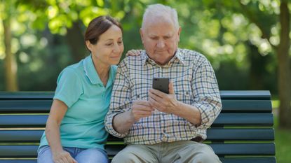 Woman sitting next to a senior man who is looking at his phone, outside on a bench in the park