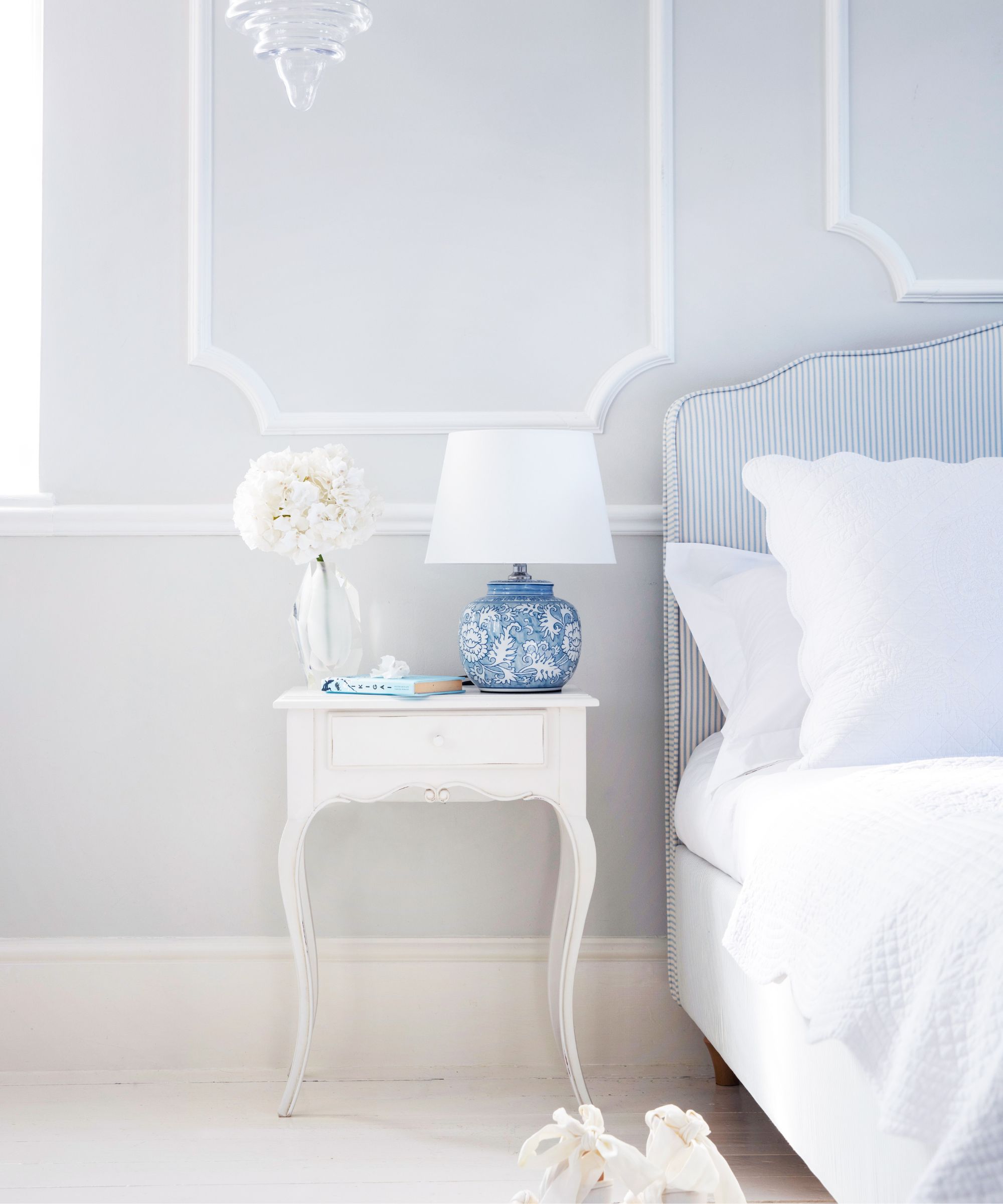 Bright and airy bedroom with a classic, elegant style, featuring a delicate white side table topped with a blue patterned lamp and fresh white flowers.