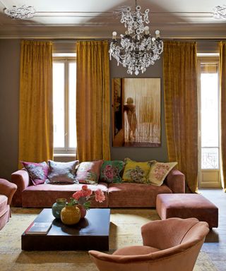 pink and ochre living room with ochre drapery, chandelier, pink modular sofa and chairs, colorful floral cushions, patterned rug and artwork of a lady