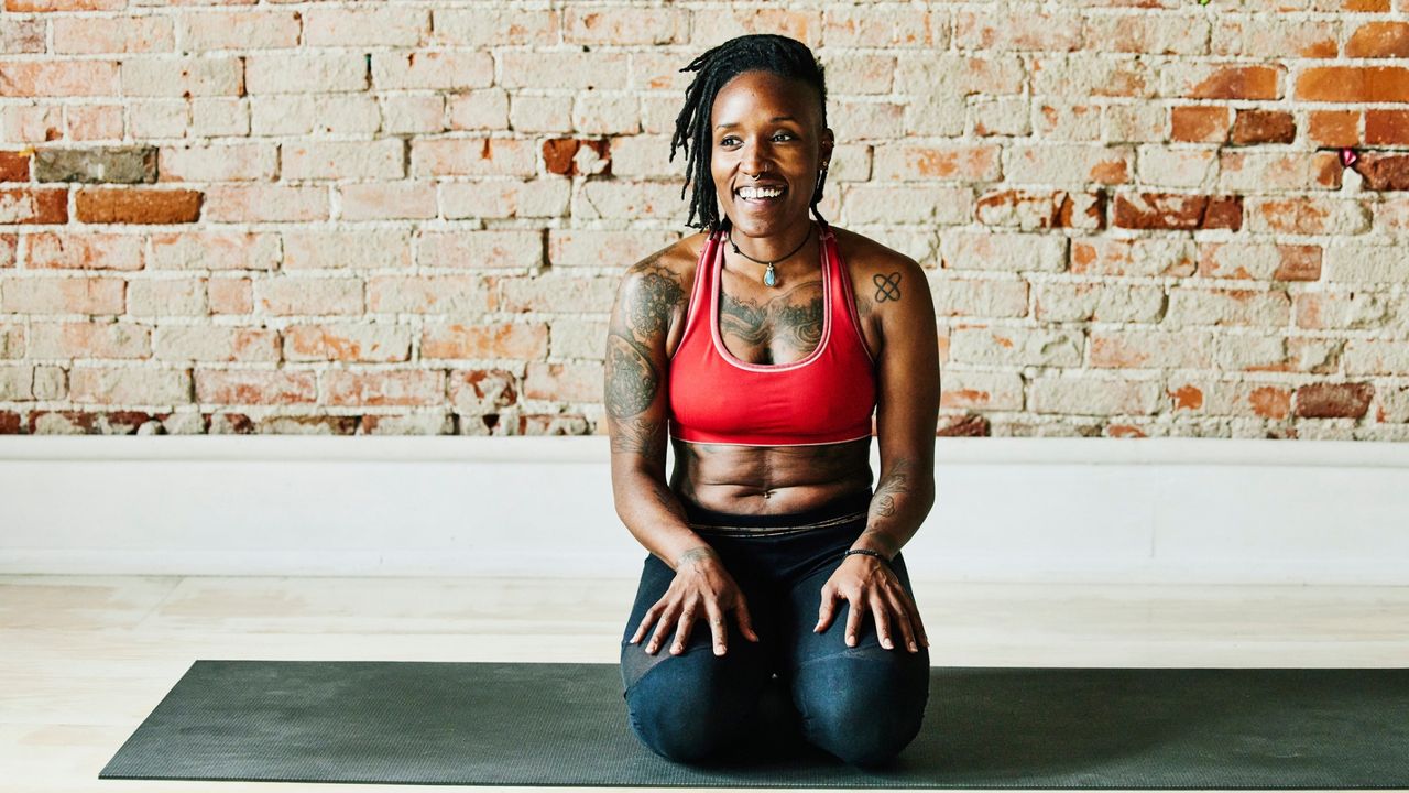 woman on a mat in a blank space with a brick wall behind her. she&#039;s wearing a red crop top and dark leggings and sat on her knees smiling