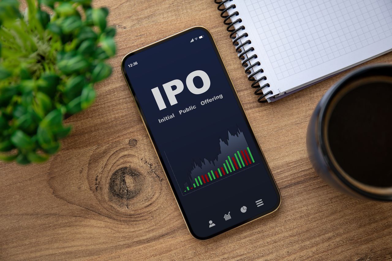 A smartphone on a table with a screen reading &quot;IPO,&quot; next to a notebook and coffee cup.