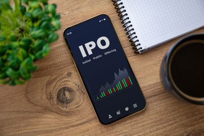 A smartphone on a table with a screen reading "IPO," next to a notebook and coffee cup.