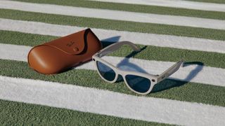 A photo of a Ray-Ban Meta smart glasses in the Skyler Shiny Chalky Gray style sitting atop a white-and-green towel next to the glasses charging case.