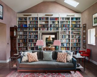 Large living room space with textured, pale pink walls, grand home library, floor to ceiling shelving design, large gray sofa with cushions, patterned rug
