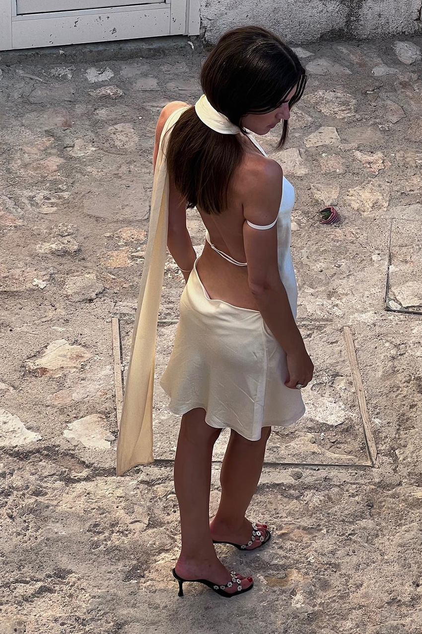 European vacation outfit idea is shown in a photo of a woman standing outside wearing a white satin mini dress with a built-in scarf and low-back styled with black heeled mules