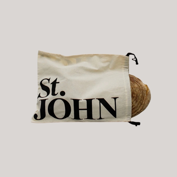 A beige bread bag with the logo "St. JOHN" in black big characters holds a loaf against a pale grey background.