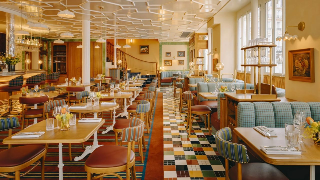 A warmly designed restaurants features a padel-shaded palette, floor tiles in different tones, wooden tables and wooden and leather chairs in mid-century modern style, and a white, plastered ceiling reflecting the light.