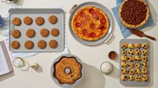 A grey set of bakeware including a cookie sheet with cookies on it, a round pizza pan with ap izza on it, a pie dish with a pecan pie inside, a muffin tray with blueberry muffins, and a bundt tin with a cake inside, one a white countertops, surroudned by baking accoutrements such as a wooden cake slice, and a blue striped dish towel. 