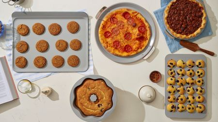 A grey set of bakeware including a cookie sheet with cookies on it, a round pizza pan with ap izza on it, a pie dish with a pecan pie inside, a muffin tray with blueberry muffins, and a bundt tin with a cake inside, one a white countertops, surroudned by baking accoutrements such as a wooden cake slice, and a blue striped dish towel. 