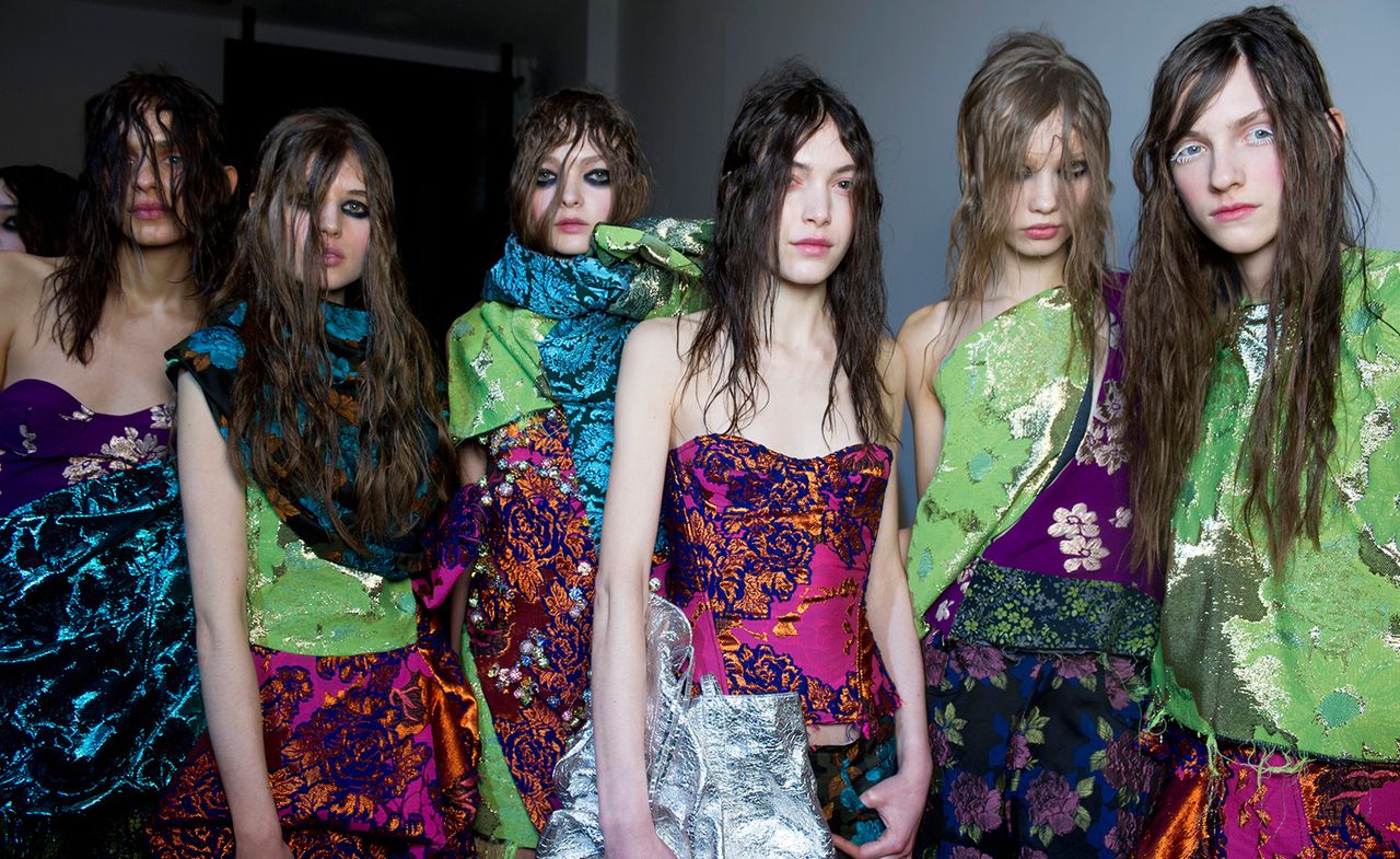 Row of six female models, dressed in colourful clothing, heavy eye makeup and messy hair styles, white wall in the backdrop