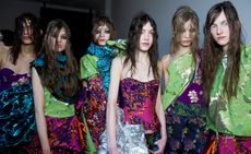 Row of six female models, dressed in colourful clothing, heavy eye makeup and messy hair styles, white wall in the backdrop