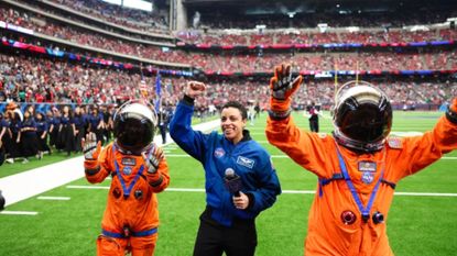 A woman with short dark hair wearing a blue suit and holding a microphone is flanked by two people wearing orange space suits with dark helmets on a football field with a full arena behind them.