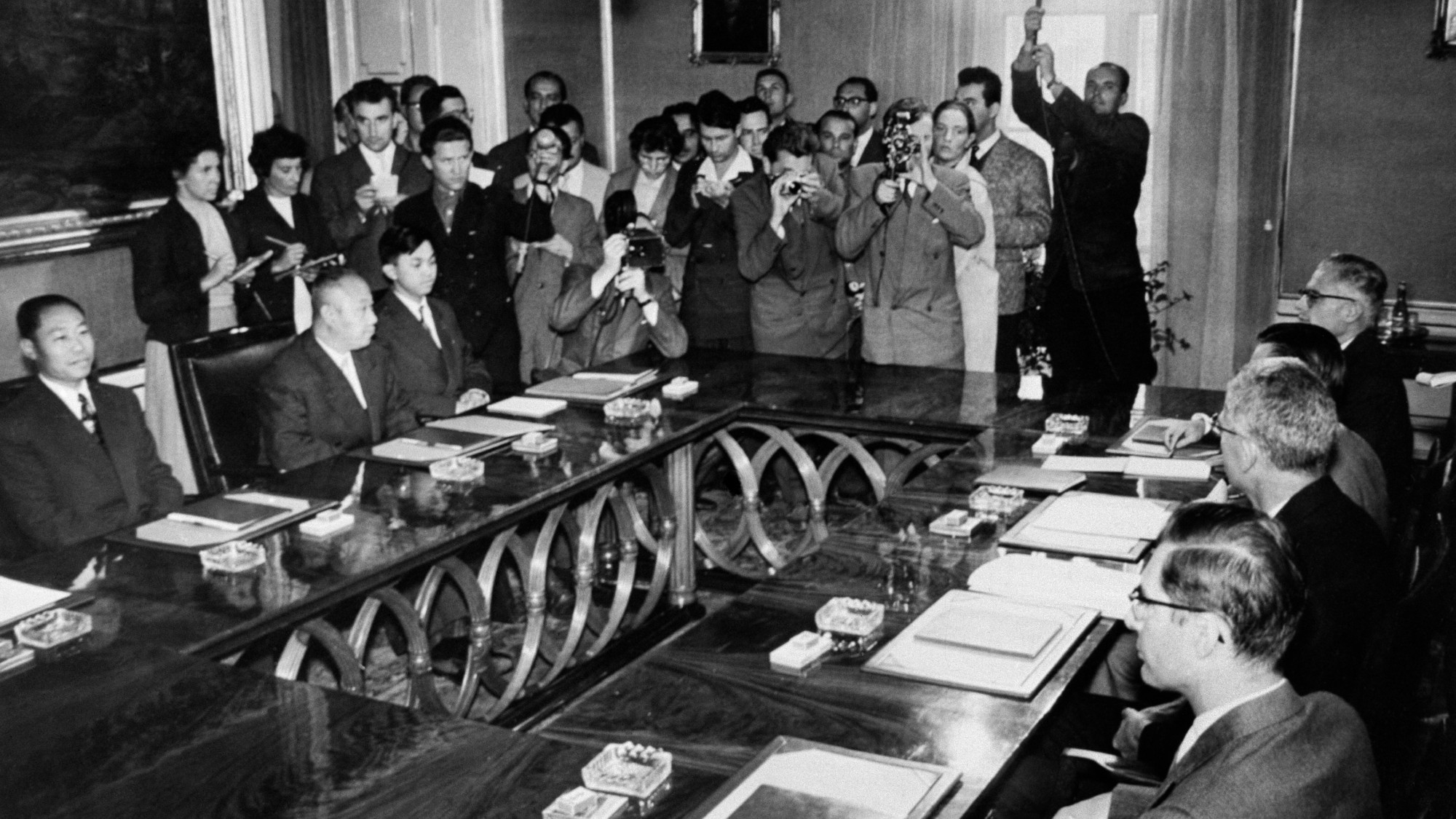 black and white photograph of a crowded meeting room with journalists, photographers surrounding two banks of tables with diplomats and officials seated