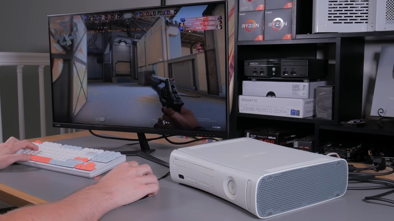 Xbox 360 with swapped internals in foreground, hands on keyboard and mouse playing Valorant on monitor in background.