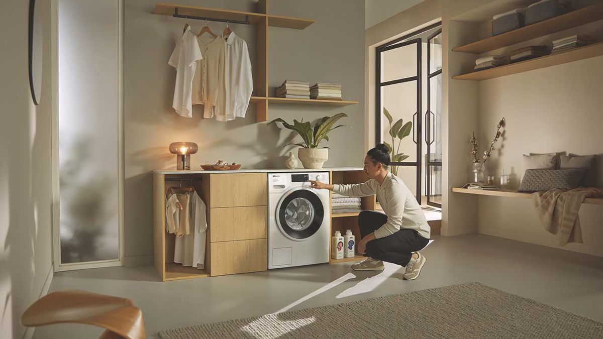 Woman bending down to load a washing machine in a bright kitchen.