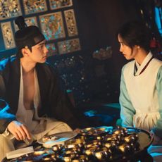 A man (Lee Chae-min) and a woman (Lim Yoon-a) wearing Joseon-era Korean clothing sit in a room in front of a table full of gold, in 'Bon Appetit, Your Majesty.'
