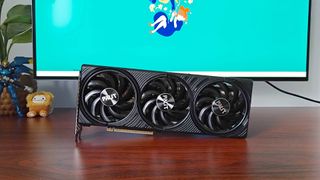 Palit GeForce RTX 5060 Ti Inifnity graphics card standing horizontally on woodgrain desk with monitor in backdrop with green wallpaper next to plant