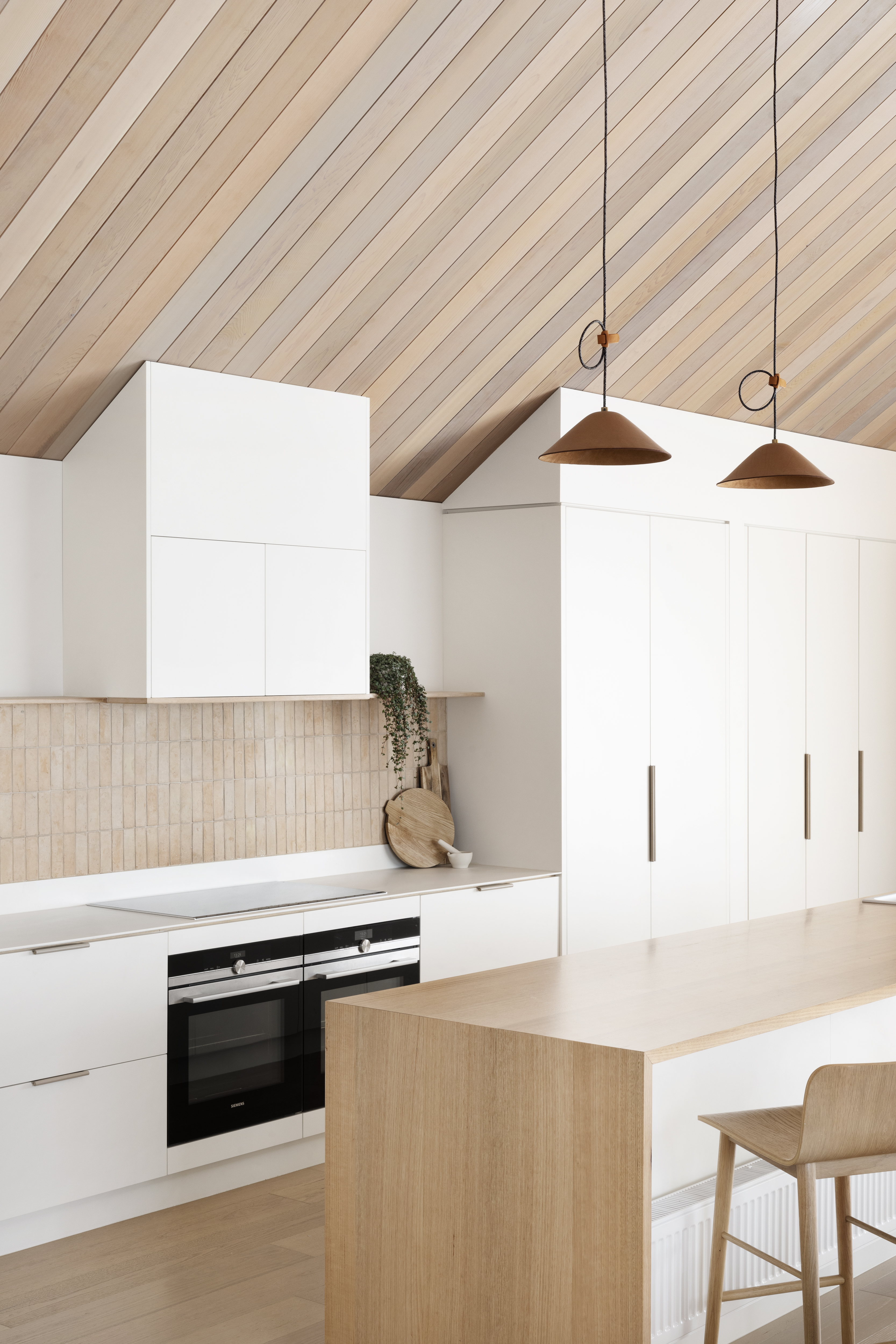 scandinavian coastal style kitchen with timber panel sloped ceilings, white cabinets, earth-toned tiled backsplash, timber and white kitchen island with timber bar stools, and two pendant lights hanging over the island
