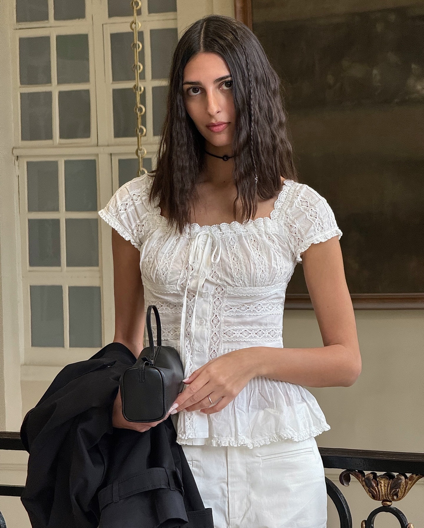 Fashion person Salome Mory shows you how to style white jeans. She wears a white pretty lace detail top, white jeans, black trench coat, black bowler bag and black and cream heels. She is looking into the camera in a recent image from an Instagram.