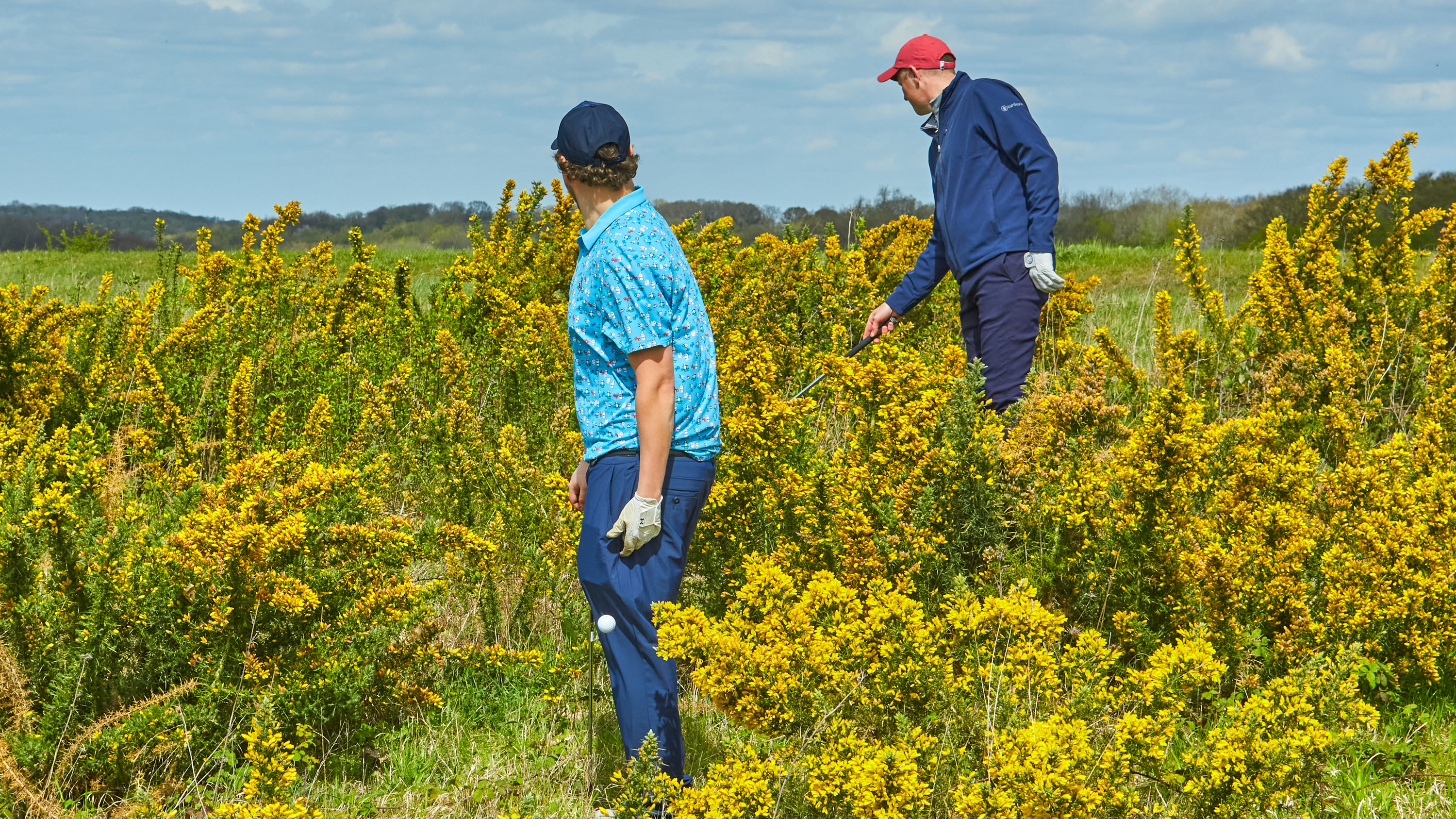 While two golfers look for a lost golf ball, one of them drops another ball behind his leg while the other carries on searching