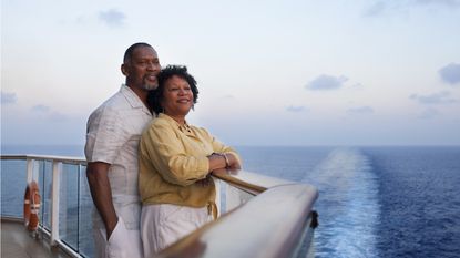 A retired couple look into the distance together while leaning on the railing of a cruise ship.