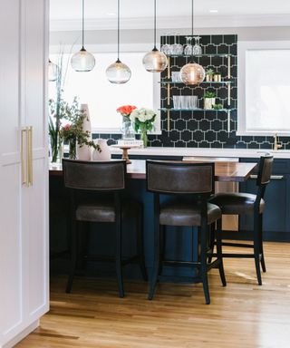 kitchen island ideas, blue and white kitchen with central island that doubles as a table, bar stools, wooden floor, tiled walls, open shelving, glass pendant lights