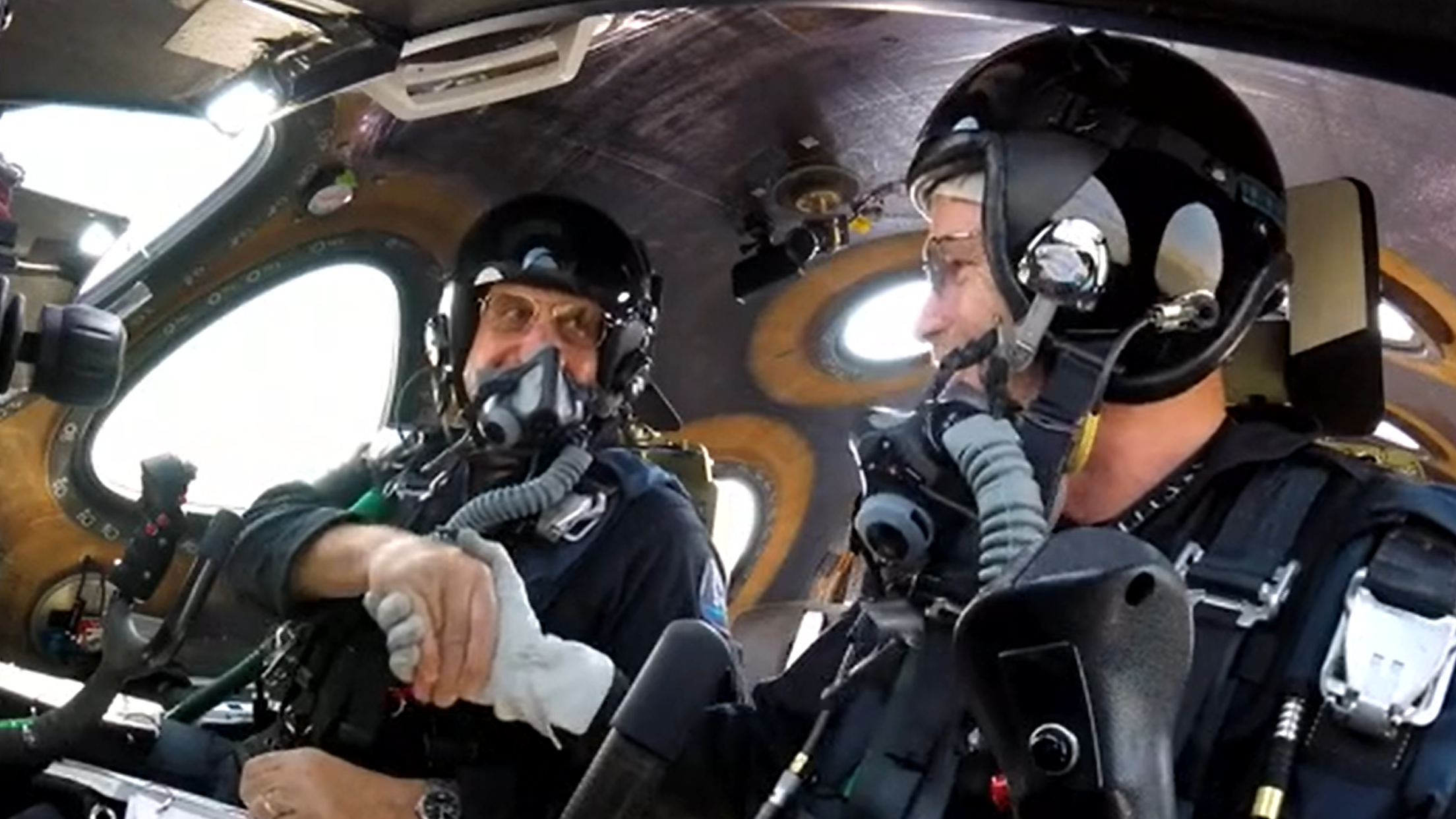 two pilots in a cockpit wearing helmets, masks and other equipment. they shake hands. windows are visible in the background