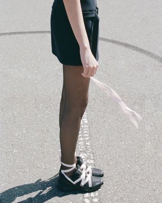 a model stands on a basketball court wearing black tights and a pair of sandy liang x salmon ballet sneakers with pink ribbons laced over the top