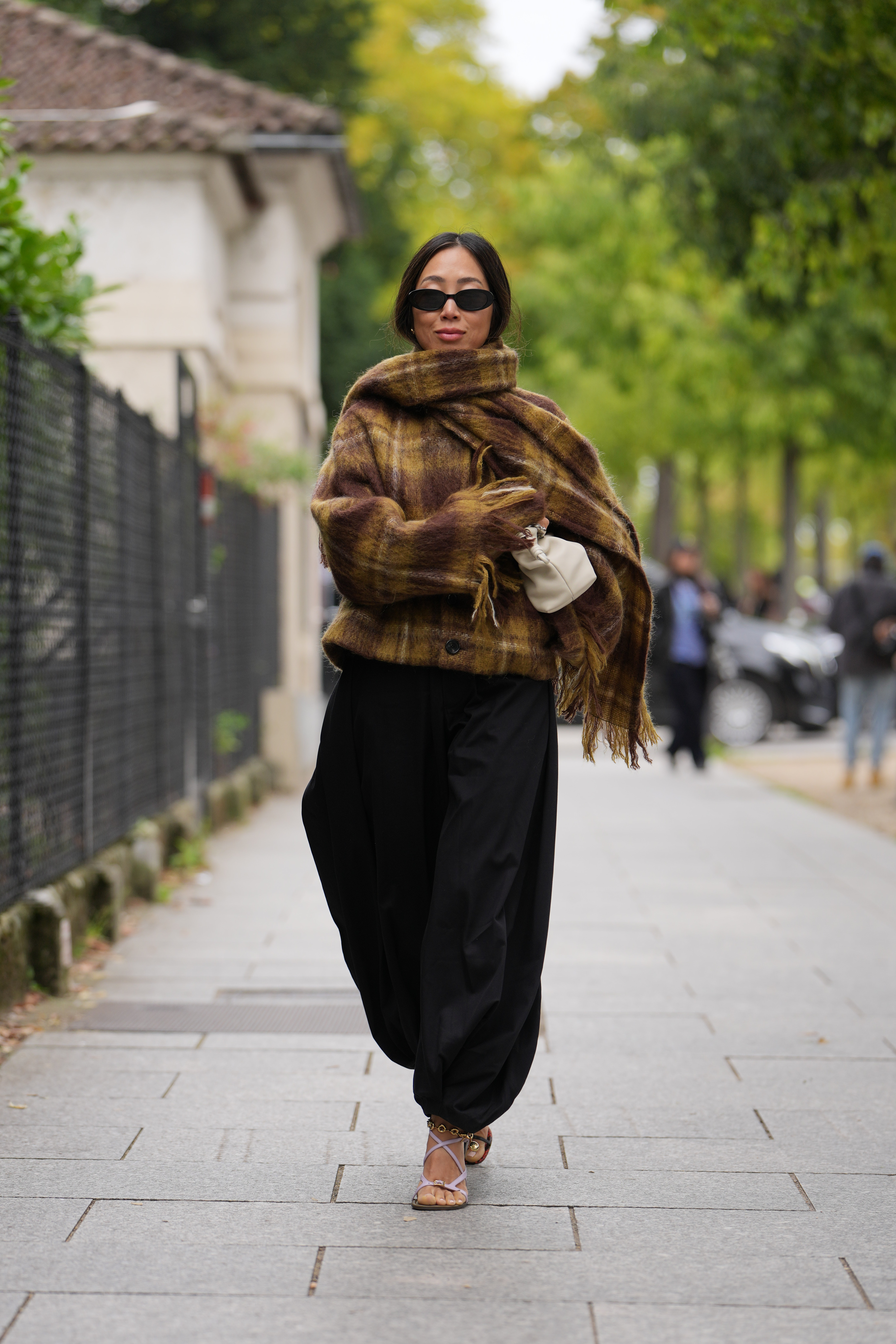 PARIS, FRANCE - SEPTEMBER 03: Aimee Song wears long dark hair parted in the center, small black oval sunglasses, silver rings and a gold chain anklet, a cream leather pouch bag, a fuzzy brown, mustard and olive plaid wool wrap with fringe layered over black voluminous gathered-ankle trousers in a matte woven fabric, tan leather strappy lace-up sandals shoes with thin straps and stiletto heels, outside Loewe, during Paris Fashion Week - Womenswear Spring Summer 2026, on September 03, 2025 in Paris, France (Photo by Edward Berthelot/Getty Images)