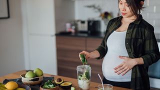 Pregnant woman putting cucumber into blender, one source of folic acid, while holding stomach